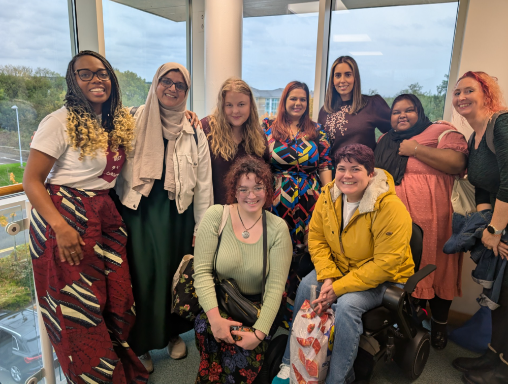 Mae naw o bobl wedi ymgynnull dan do ger ffenestri mawr yn nigwyddiad Cysters. Mae un person yn eistedd mewn cadair olwyn, gyda rhywun arall yn hwtian wrth ei hymyl. Mae pawb yn gwisgo dillad lliwgar â phatrymau, ac mae rhai yn gwisgo hijab. Mae pawb yn edrych tuag at y camera.
