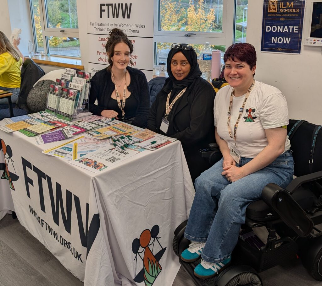 Mae Rachel, Noward, a Dee yn gwenu ar y camera wrth iddynt eistedd y tu ôl i fwrdd arddangos ar gyfer FTWW (Triniaeth Deg i Fenywod Cymru) ac wrth ei ochr. Mae’r bwrdd wedi’i orchuddio â lliain gwyn gyda’r logo FTWW arno. Ar y bwrdd, ceir amrywiaeth o ddeunyddiau lliwgar wedi’u hargraffu fel taflenni, pamffledi, a phennau ysgrifennu wedi’u trefnu’n daclus. Y tu ôl i’r bwrdd, mae baner yn arddangos yr enw FTWW a thestun dwyieithiog yn Gymraeg a Saesneg.