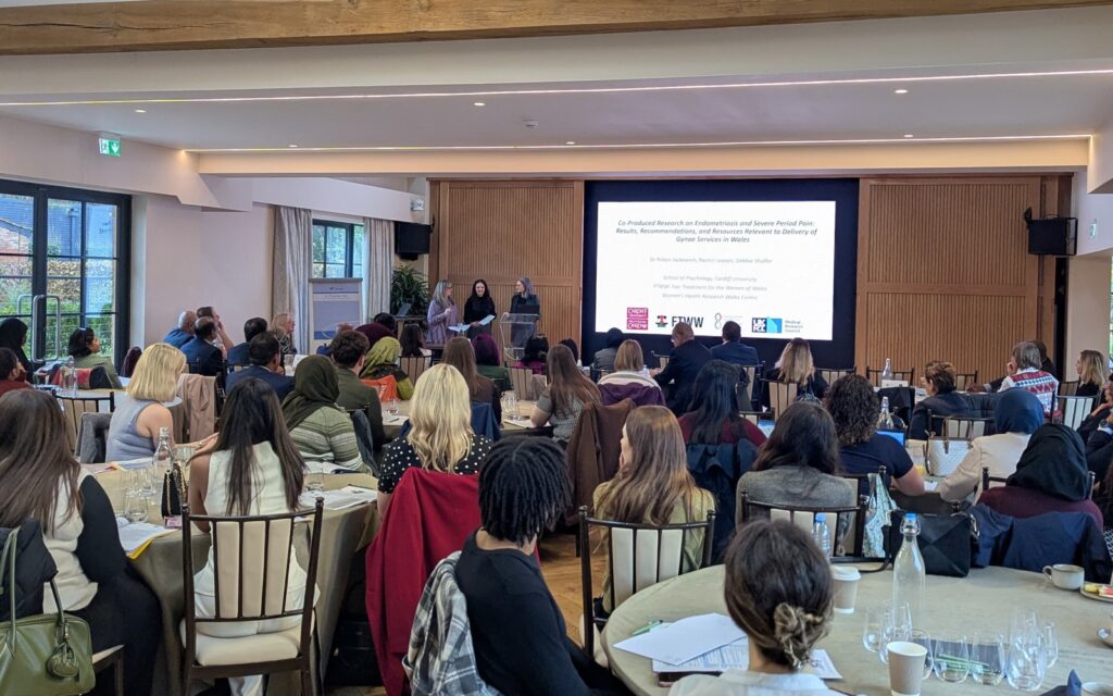 Conference room with attendees seated at round tables facing a large screen. Debbie, Rachel, and Robyn stand at the front. The presentation slide reads: “Co-produced Research on Endometriosis and Severe Period Pain: Results, Recommendations, and Resources Relevant to Delivery of Gynae Services in Wales” with logos for Cardiff University, FTWW, and partners.