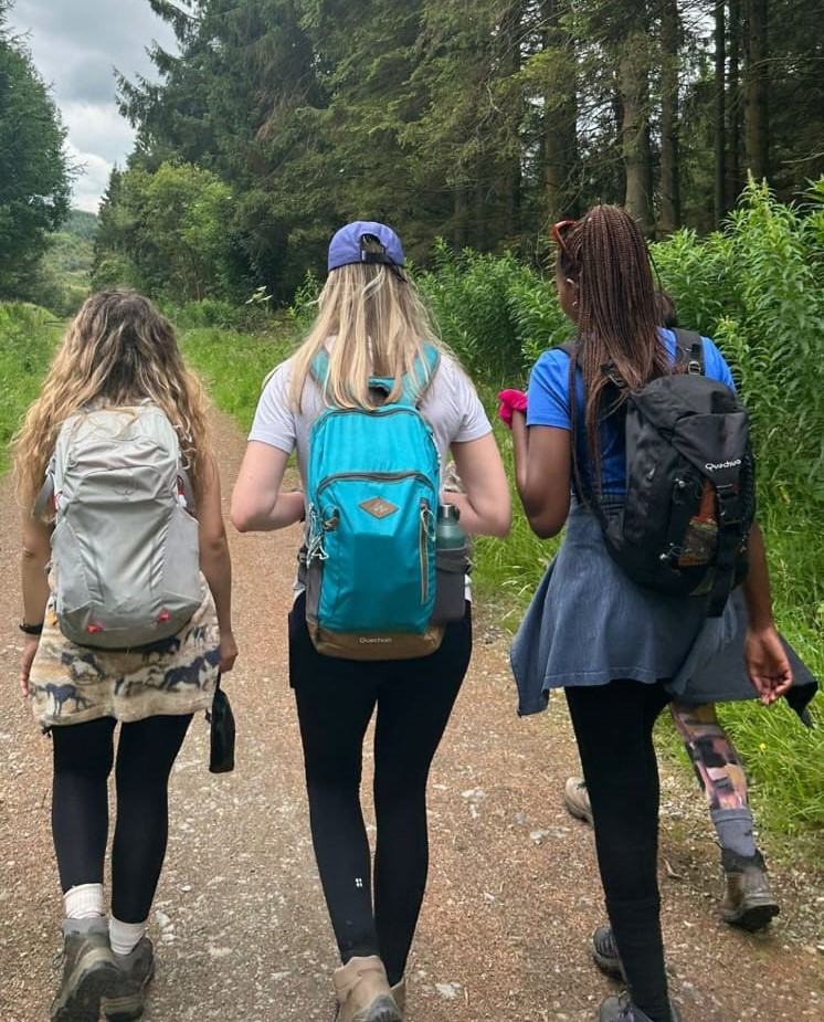 Four women walking along a forest trail, each carrying a backpack, surrounded by tall trees and greenery