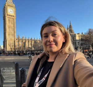 Menyw â gwallt golau yn gwisgo côt lliw brown golau gyda laniard gwirfoddolwr FTWW yn sefyll wrth ymyl Big Ben a’r Tŷ Cyffredin