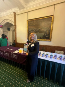 Menyw â gwallt golau yn gwisgo dillad du gyda laniard gwirfoddolwr FTWW  yn sefyll o flaen llun yn y Tŷ Cyffredin. Mae’n gwenu ar y camera ac yn dal adroddiad ‘Mothers for Mothers’ yn ei dwylo.