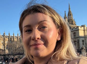 A woman, who is Eliza, wearing a beige coat and an FTWW lanyard smiles while standing in front of the Houses of Parliament.