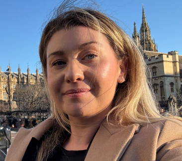 A woman, who is Eliza, wearing a beige coat and an FTWW lanyard smiles while standing in front of the Houses of Parliament.