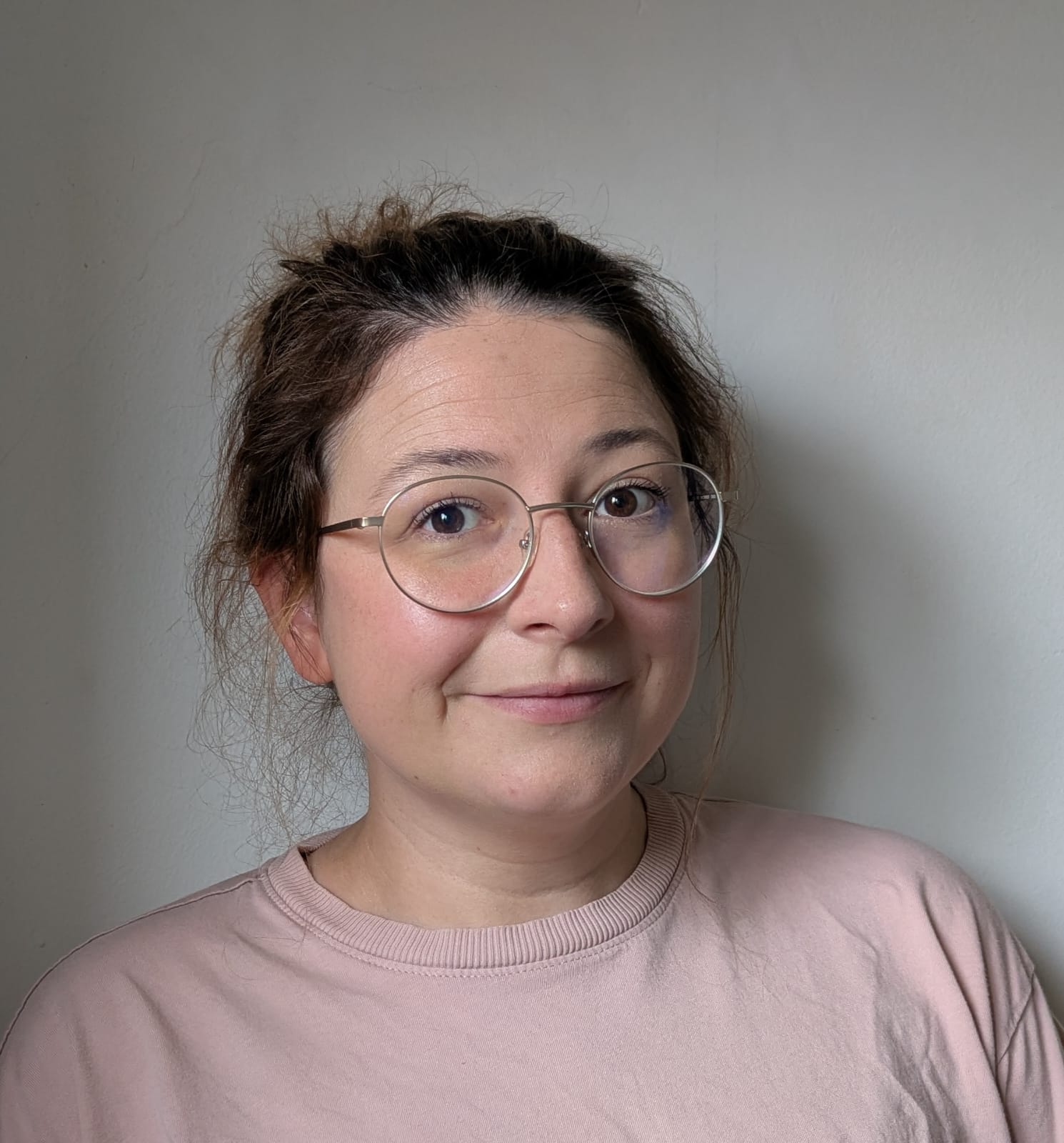 A picture of Isabel, wearing a pink top and glasses smiling in front of a grey background.